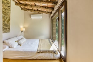 Une chambre avec un mur en pierre, un plafond en bois, un appareil de climatisation, un lit avec du linge de lit blanc, une lampe de chevet et une vue par la fenêtre.