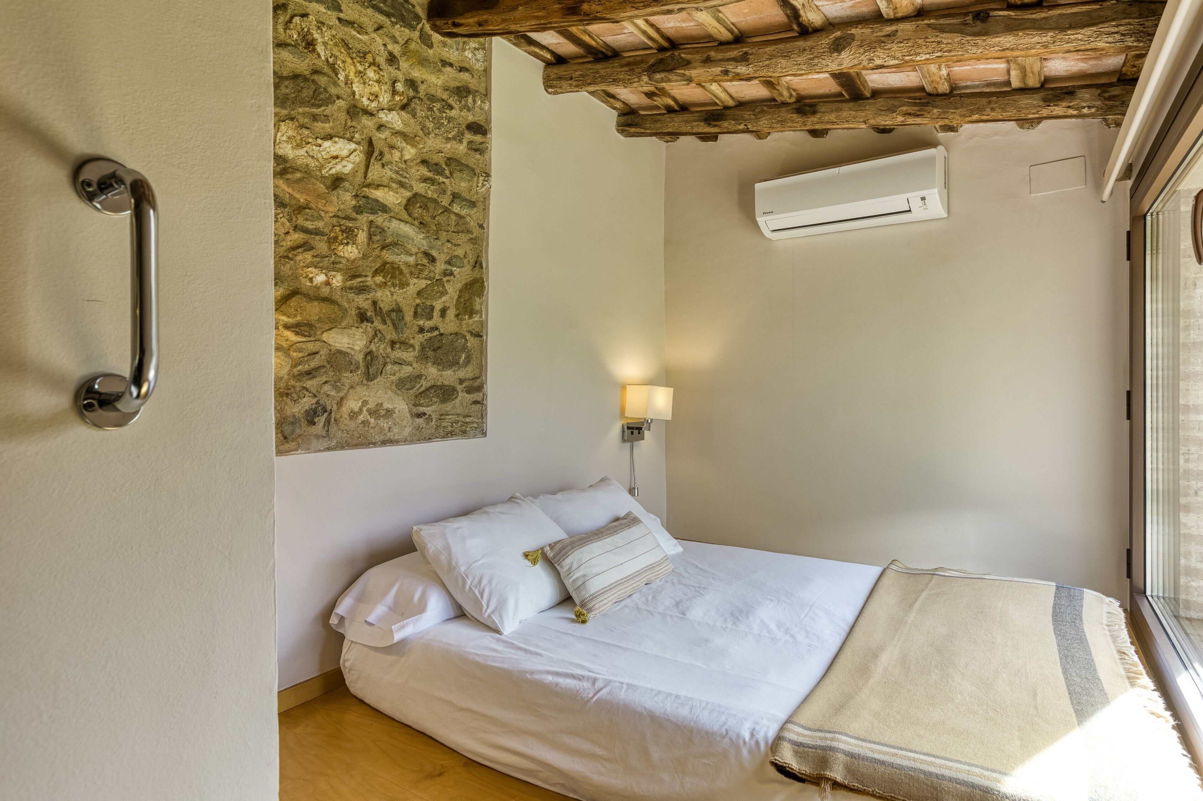 Un dormitorio con pared de piedra, una cama con ropa de cama blanca, techo de madera, aire acondicionado y una mesita de noche con lámpara.
