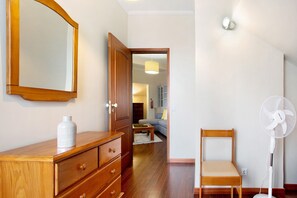 A room with a wooden dresser, a white vase, a wooden chair, a standing fan, and a mirror on the wall.