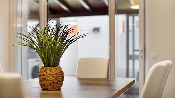 Una habitación con una mesa de madera, una maceta tejida, una silla blanca y un ventanal de vidrio que da a un espacio exterior.