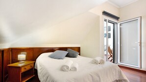 Un dormitorio con cabecera de madera, mesita de noche con lámpara, cama con ropa de cama blanca, dos almohadas y un juego de toallas.