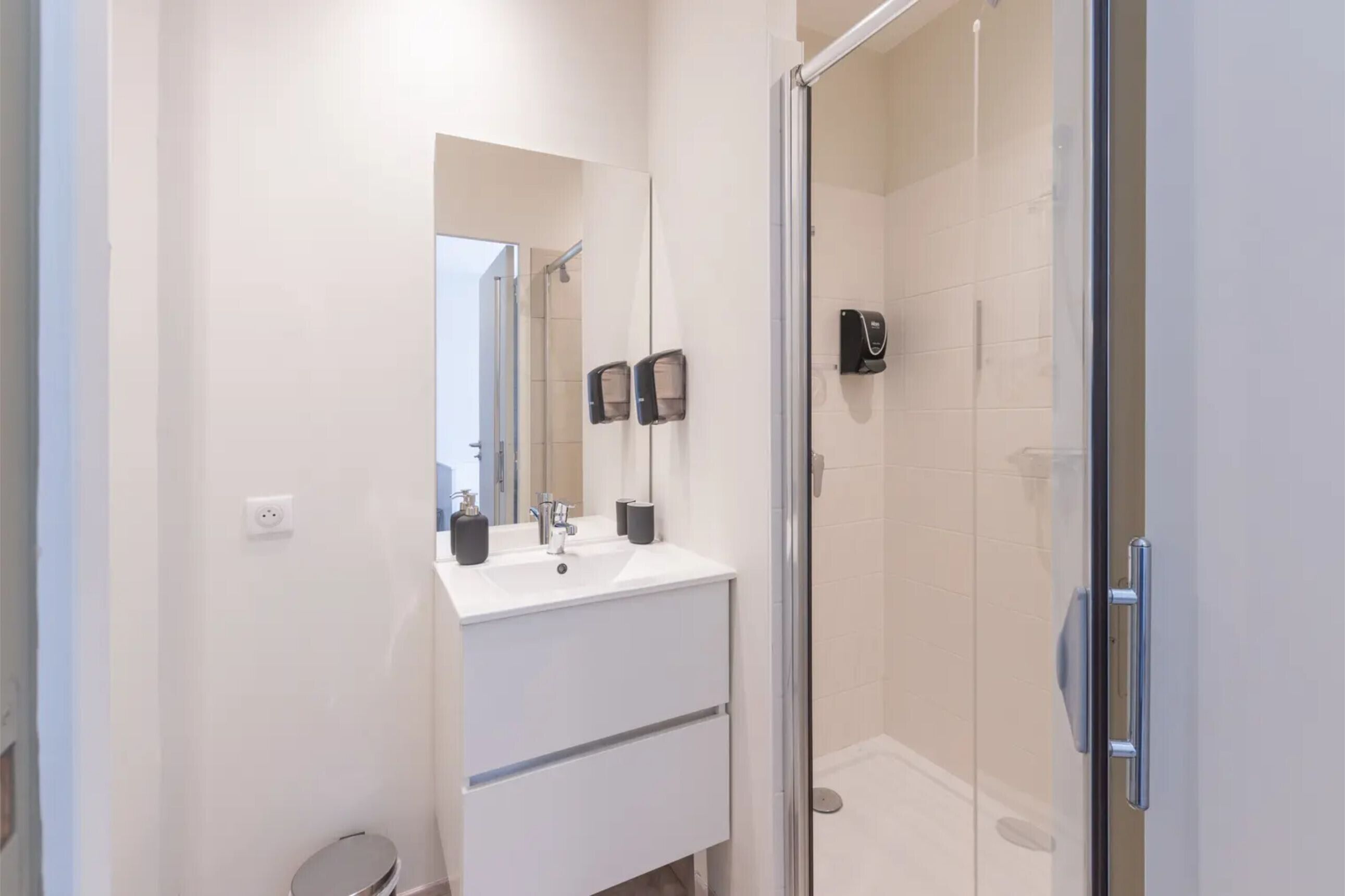 A modern bathroom with a glass shower enclosure, a white sink with a mirror, and two black soap dispensers.