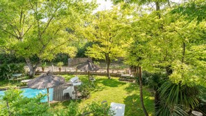 Zona de piscina amb una ombra de palla, seients de relaxació blancs i un edifici petit, tot envoltat de vegetació abundant i arbres.