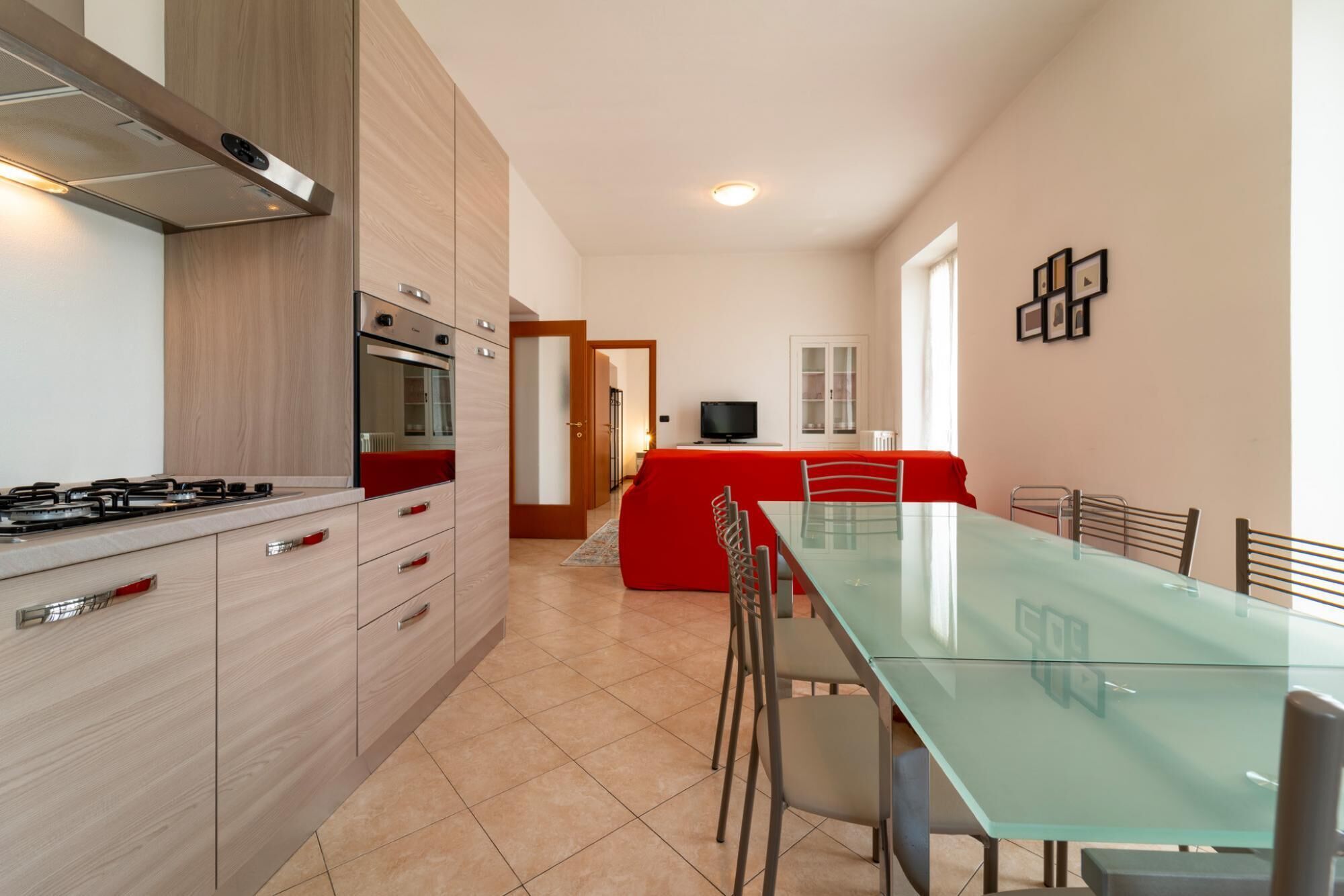 A modern kitchen with a gas stove, oven, and microwave. There is a dining area with a glass table and chairs, and a red sofa.