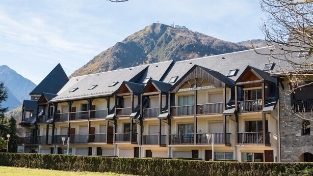 Un hotel de varios pisos con balcones, un paisaje montañoso al fondo y un seto en primer plano.