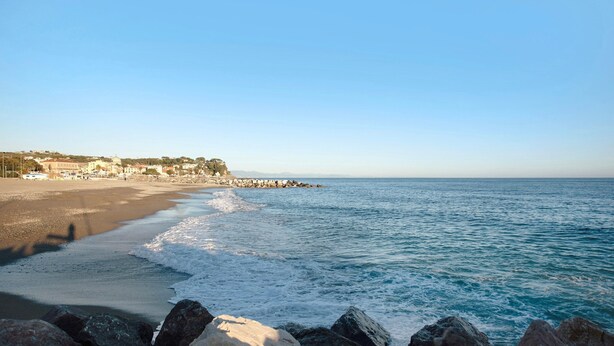 Une plage avec un premier plan rocheux, une mer calme et des bâtiments en arrière-plan.