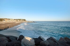 Une plage avec un premier plan rocheux, une mer calme et des bâtiments en arrière-plan.