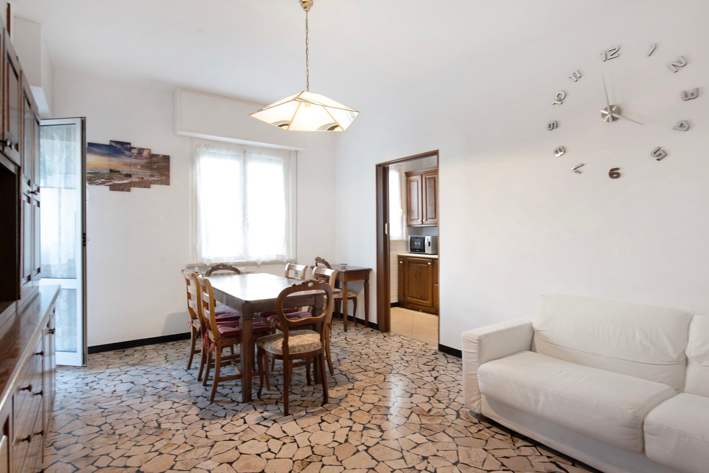 A dining area with a table and chairs, a white sofa, a clock on the wall, and a kitchen in the background.