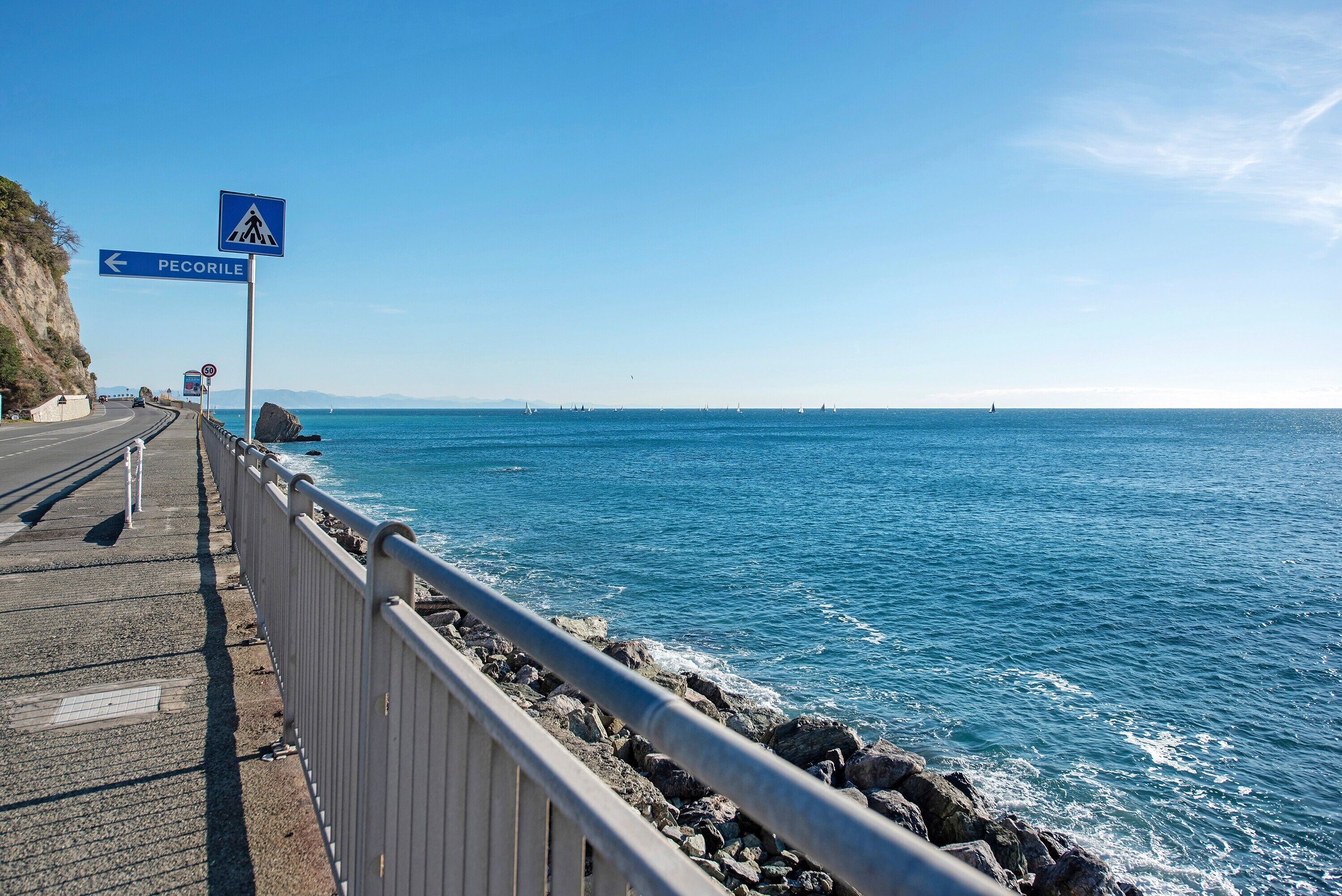 Una carretera costanera amb un senyal de pas de vianants, un indicador de direcció cap a PECORILE i un litoral rocos.