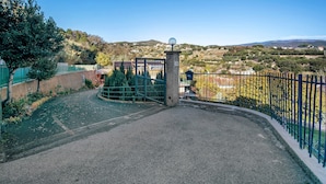 Ein verschlossener Eingang mit einem Metallgitter, einem befestigten Weg und Blick auf eine hügelige Landschaft mit Bäumen und Gebäuden.