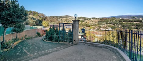 A gated entrance with a metal gate, a paved path, and a view of a hilly landscape with trees and buildings.