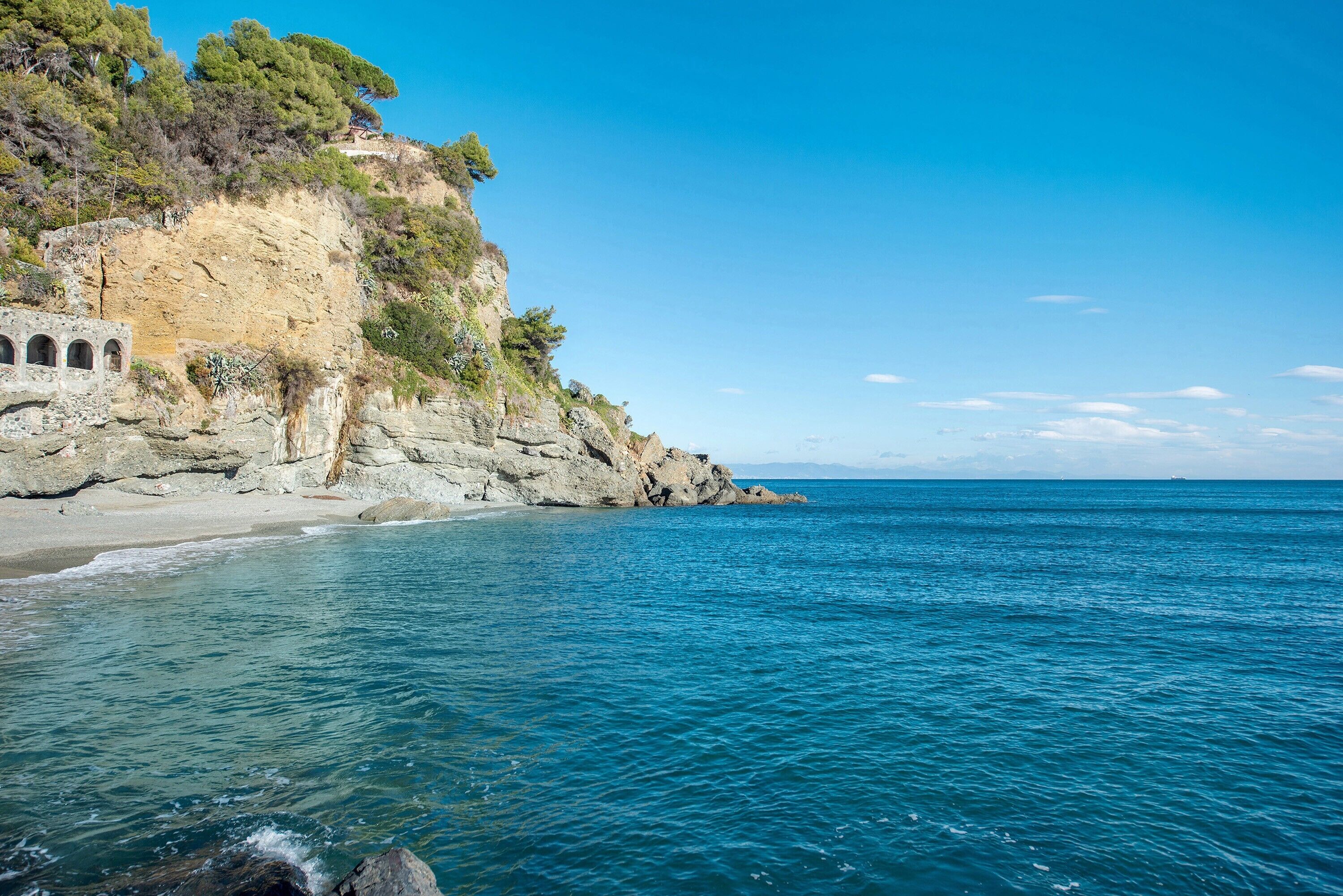 A coastal landscape with a rocky cliff, clear blue sea, and a stone structure with arches.