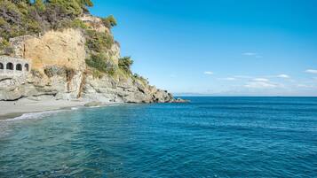 A coastal landscape with a rocky cliff, clear blue sea, and a stone structure with arches.