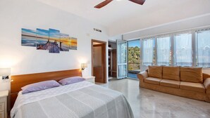 Un dormitorio con una cama, cabecera de madera, ventilador de techo, una ventana grande y un balcón.
