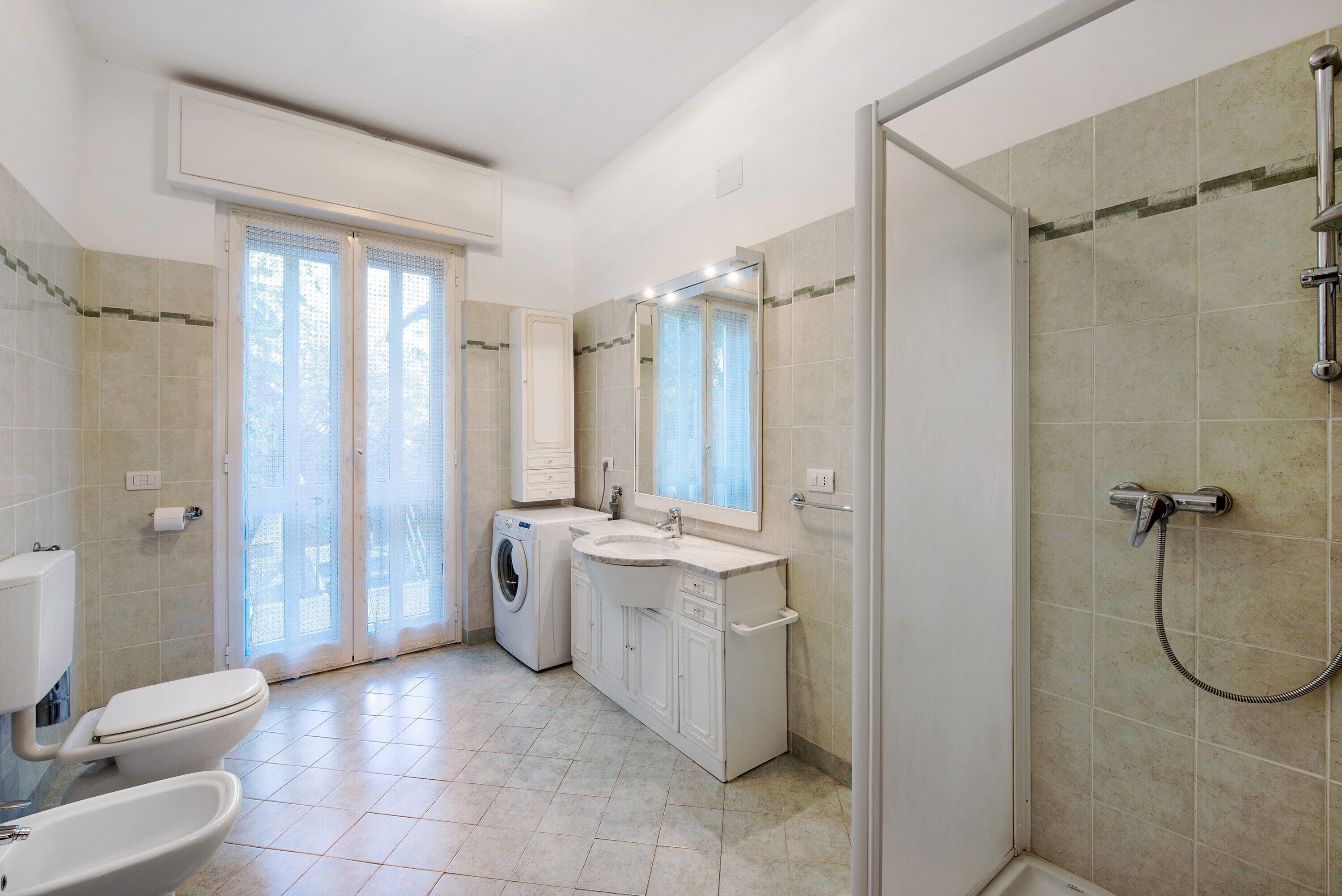 Une salle de bain comprenant des toilettes, un bidet, un lave-linge, un lavabo, un miroir, une douche et des murs carrelés.
