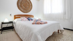 Una cama bien hecha con edredón blanco, cabecera de madera, tapiz de pared tejido y mesita de noche con lámpara.