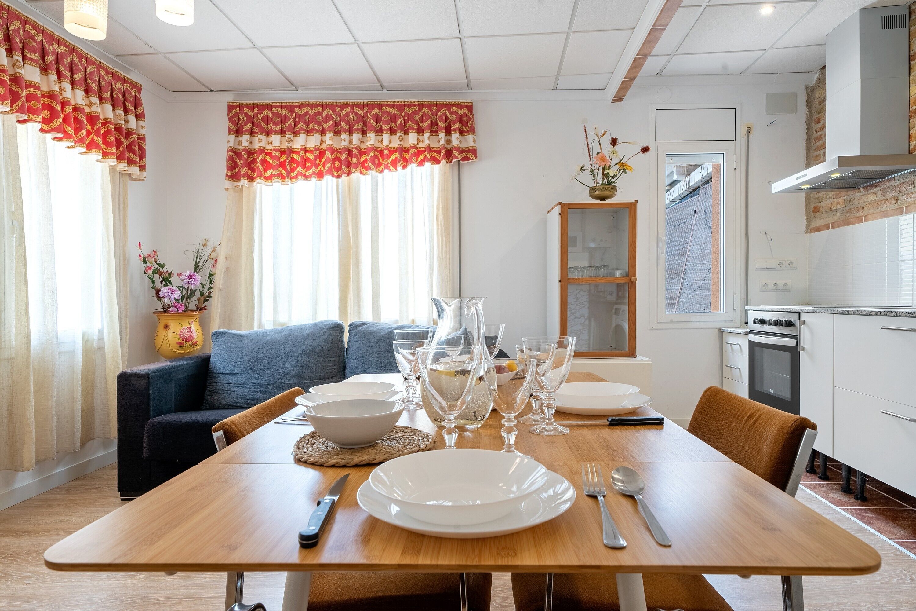 Un comedor con una mesa de madera dispuesta para cuatro personas, un sofá y una cocina al fondo.