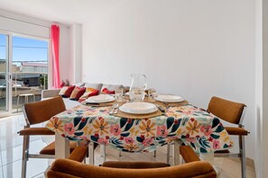 Uma sala de jantar com uma mesa posta para quatro pessoas, cadeiras, uma janela com cortina rosa e vista para edifícios.