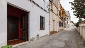 A narrow street with buildings on either side, a potted plant on the left, and a door with a red interior.