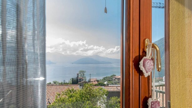 Une vue depuis une fenêtre dotée d’une porte en bois et d’un rideau, donnant sur un paysage côtier avec des montagnes et une étendue d’eau.