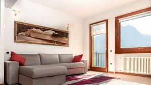 Un salon avec un canapé gris, un grand tableau représentant un paysage, une porte en bois donnant sur un balcon et une vue sur les montagnes.