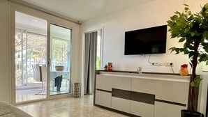 Un salon moderne avec un téléviseur à écran plat fixé au mur, une grande plante en pot et une porte-fenêtre coulissante donnant sur un espace extérieur.