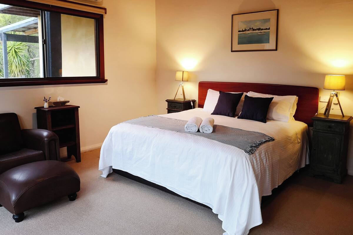 Een net opgemaakt bed met witte lakens en twee opgerolde handdoeken, een fauteuil van bruin leer, een klein houten nachtkastje en een lijst met een afbeelding aan de muur.