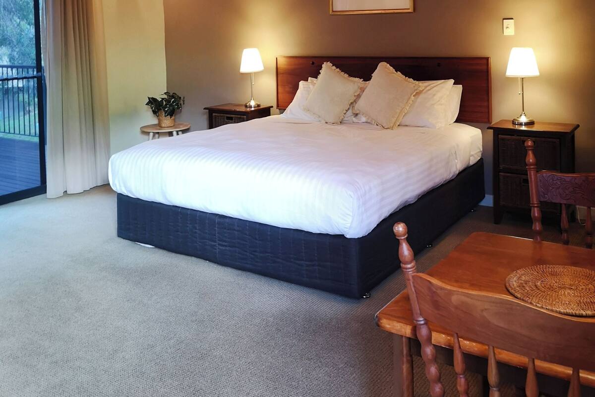 Een hotelkamer met een groot bed, twee nachtkastjes, een houten hoofdbord, een schilderij aan de muur en een balkon met gordijnen.