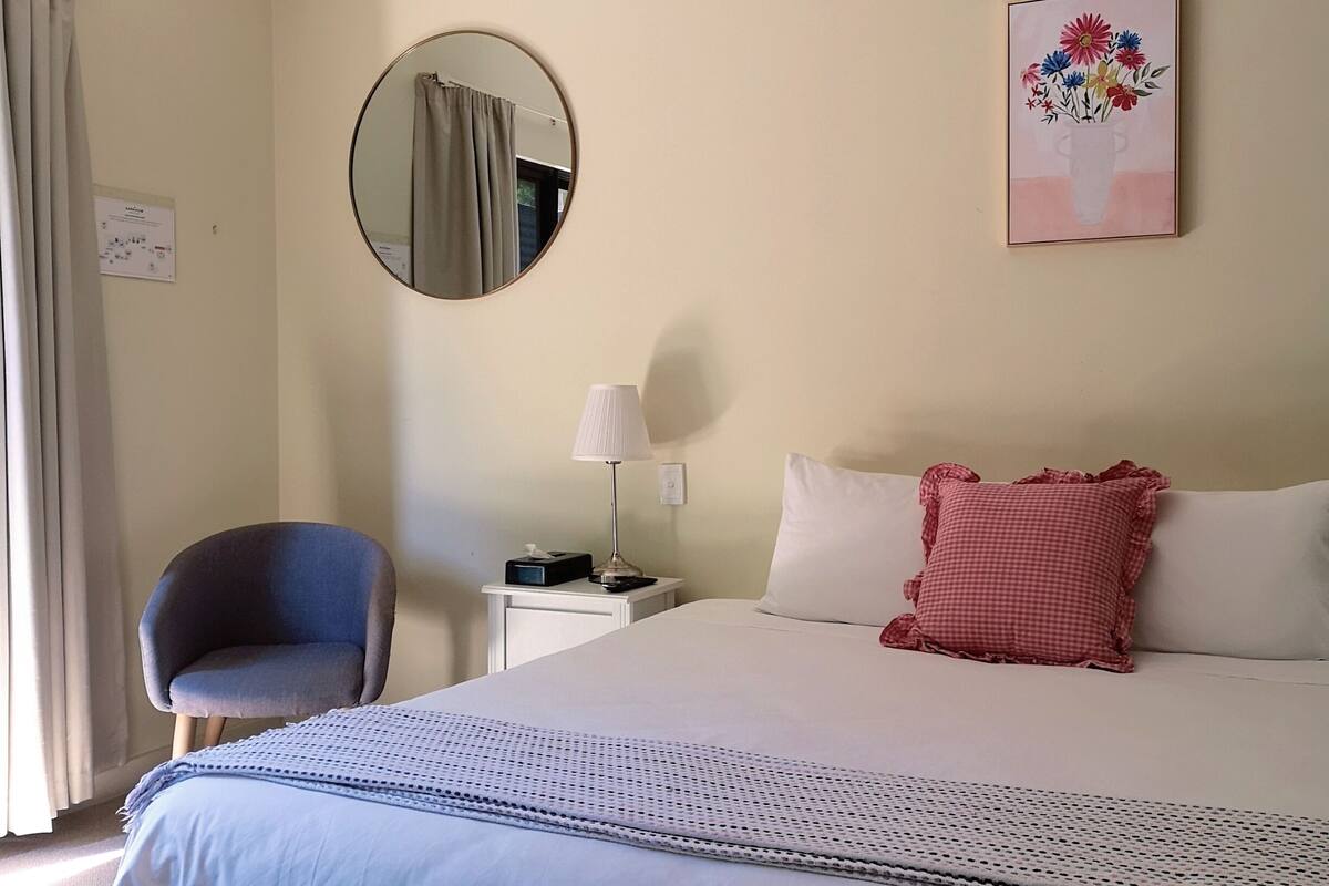 Een hotelkamer met een bed, een stoel, een nachtkastje met een lamp, een spiegel en een schilderij aan de muur.