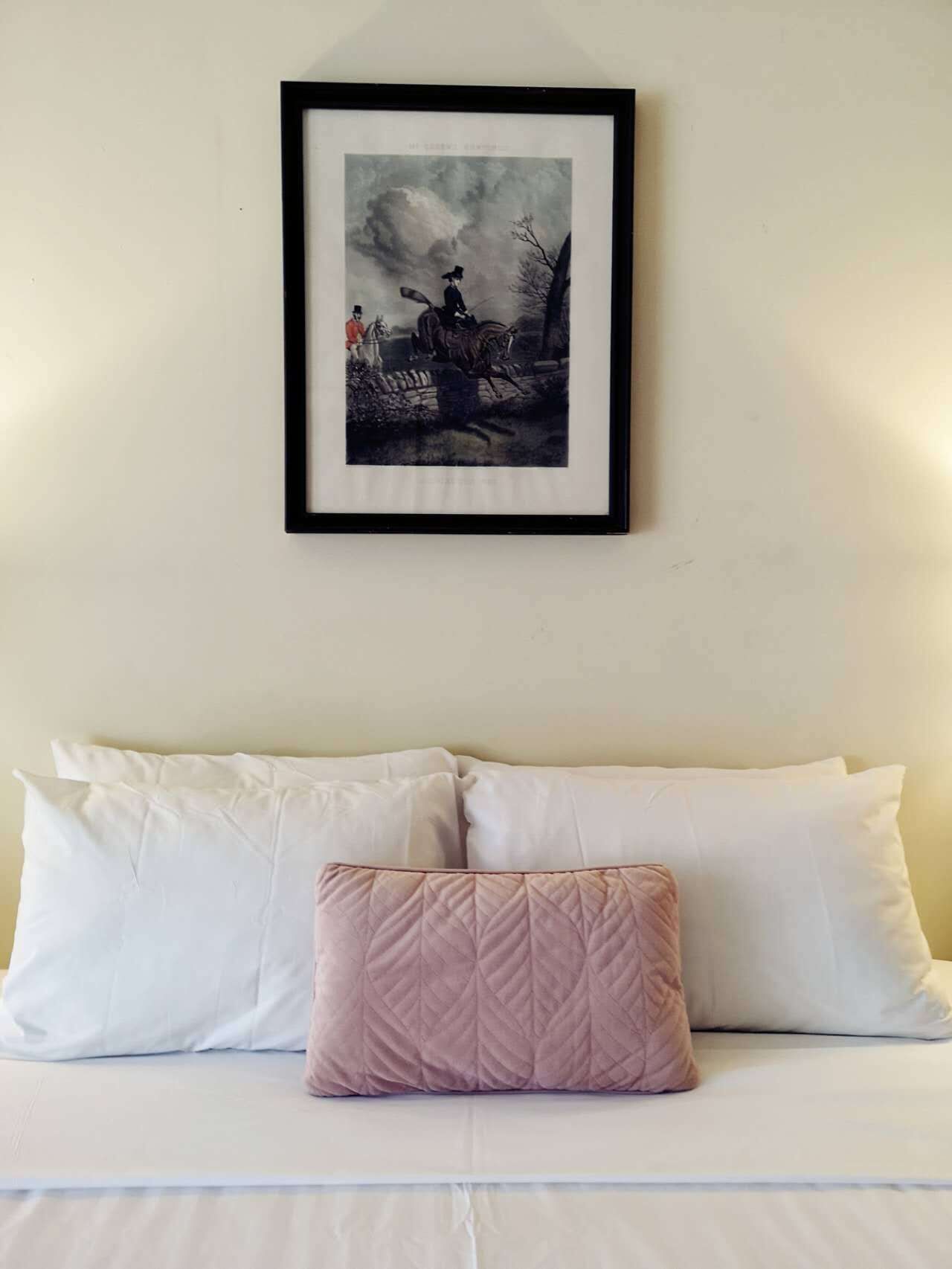 A bed with white linens and a single pink quilted pillow. Above the bed, there is a framed picture of a historical scene with two figures on horseback.