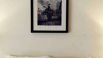 A bed with white linens and a single pink quilted pillow. Above the bed, there is a framed picture of a historical scene with two figures on horseback.