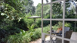 A patio with a table and chairs, surrounded by lush greenery and a view through a glass door.
