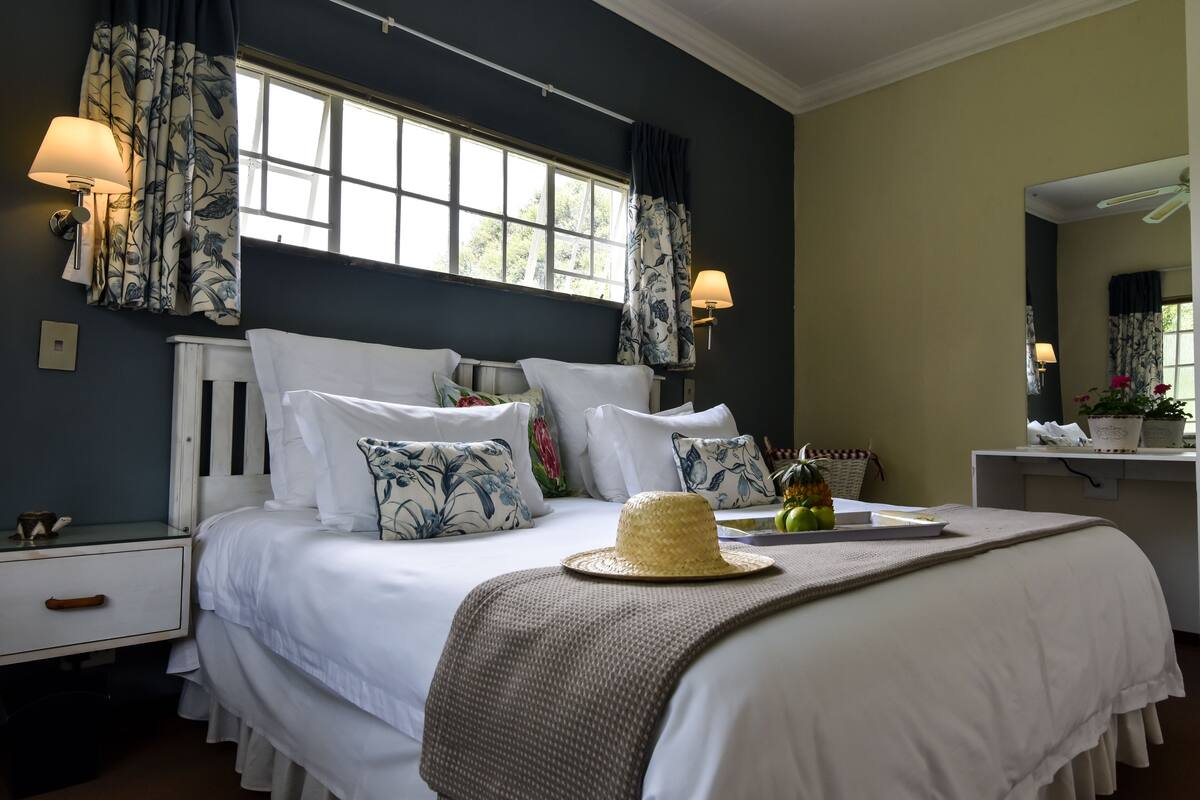 A neatly made bed with white linens, a straw hat, and a tray with fruits on a bedside table.