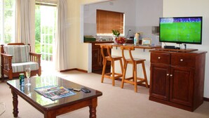A living room with a glass coffee table, wooden chairs, a TV, and a kitchen area with a window.