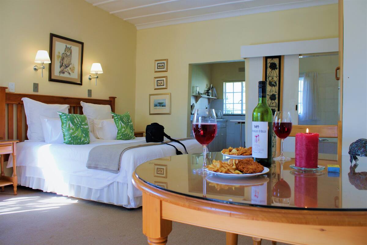 A hotel room with a bed, a dining table with food and drinks, and a kitchen area visible through an open door.