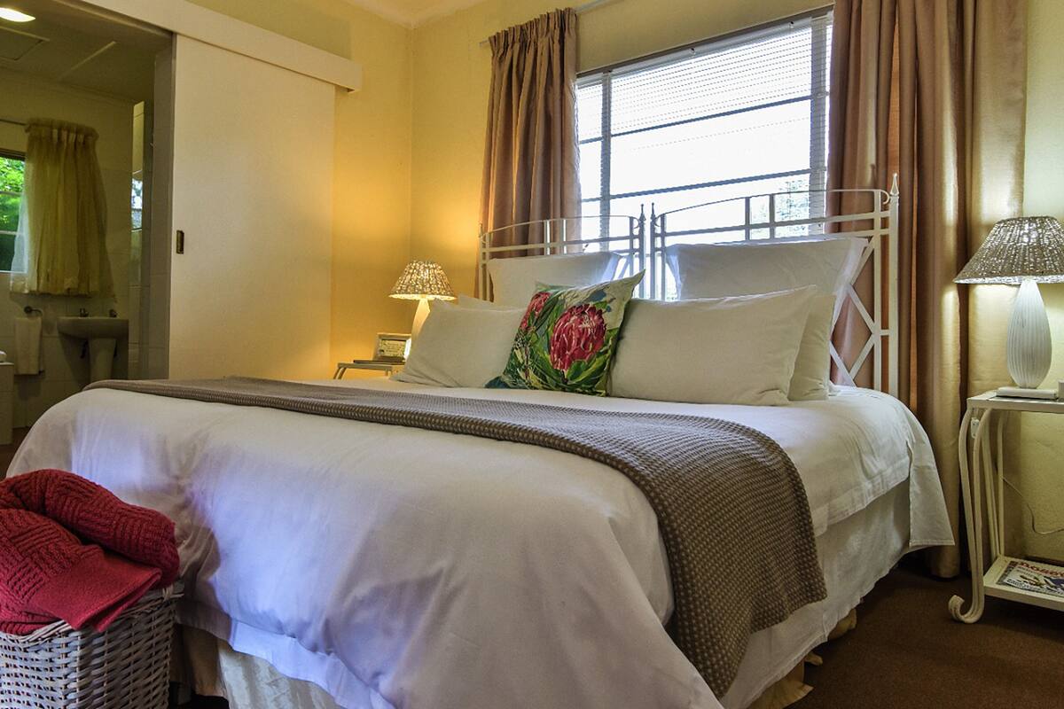 A hotel room with a bed, a nightstand, a lamp, a window with curtains, and a bathroom visible in the background.