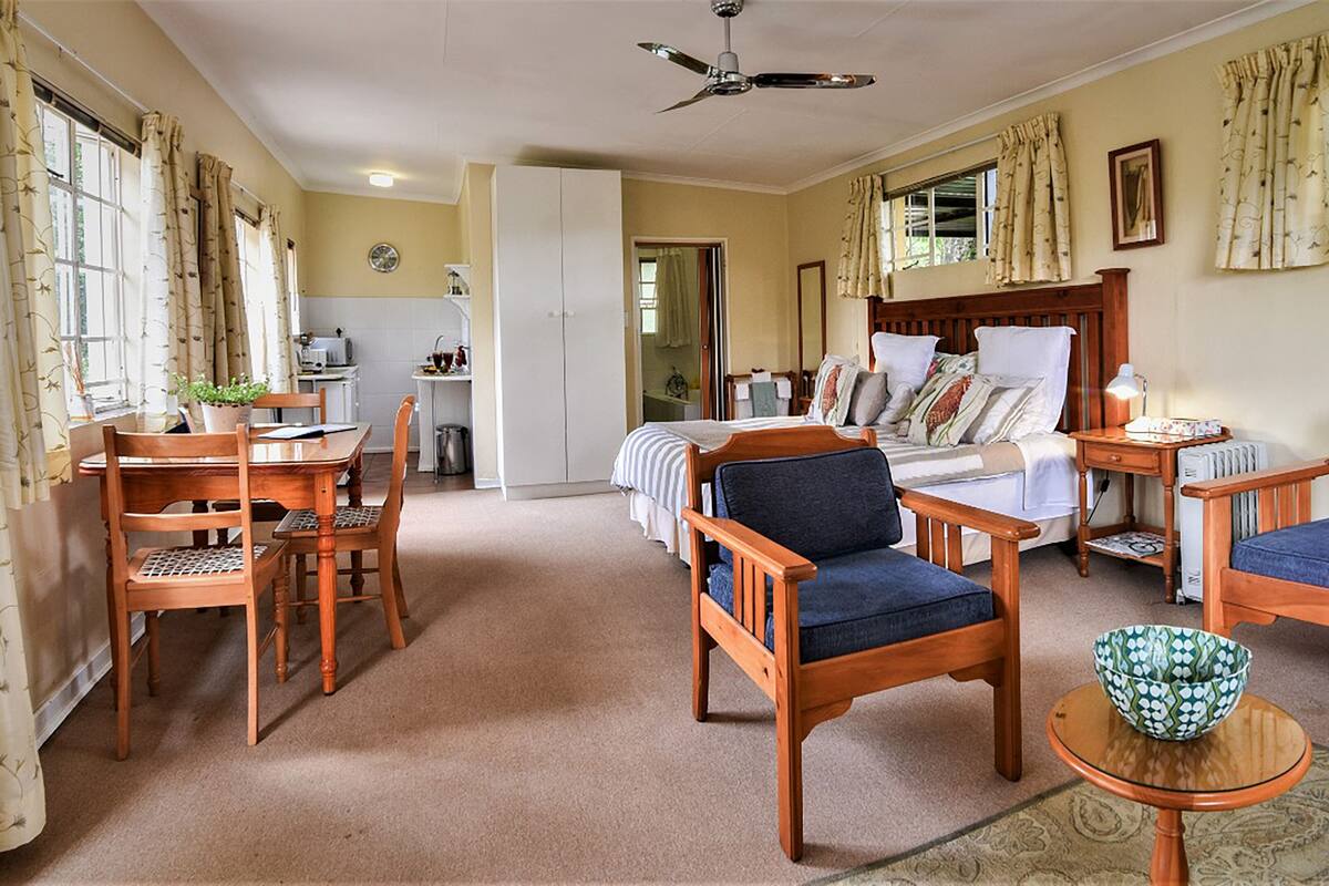 A cozy room with a bed, two wooden chairs, a table with a bowl, and a ceiling fan.