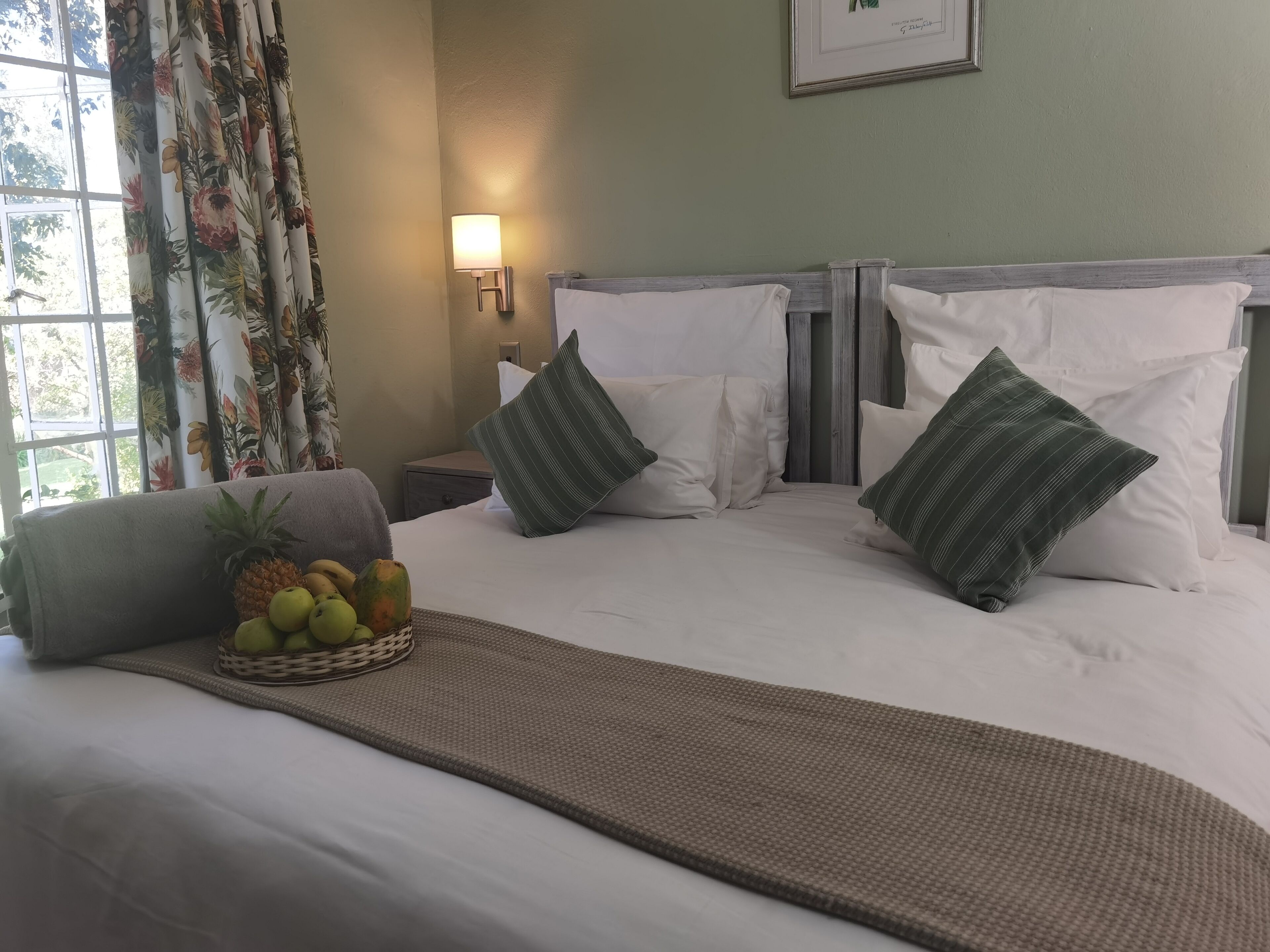 A neatly made bed with white linens and green striped pillows, a woven mat, and a fruit basket on the side.