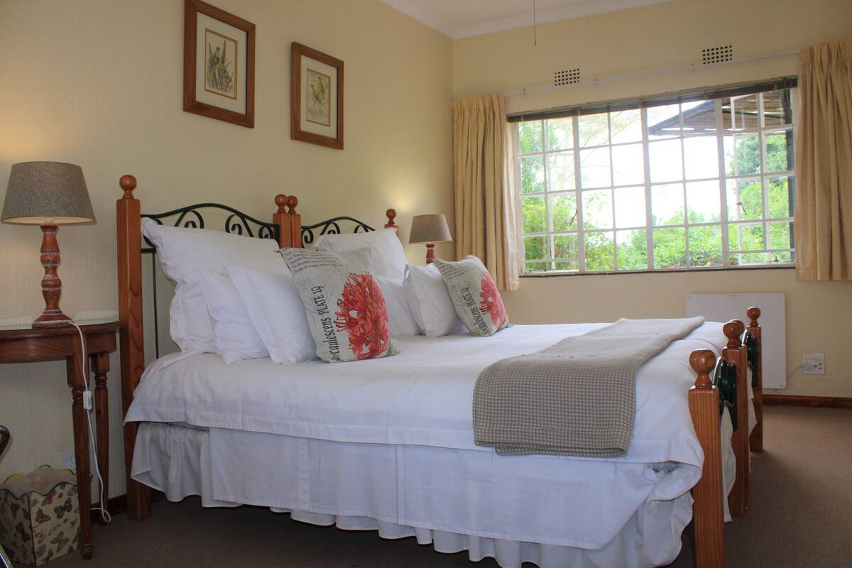A neatly made bed with white linens and decorative pillows, a bedside table with a lamp, and a window with curtains.
