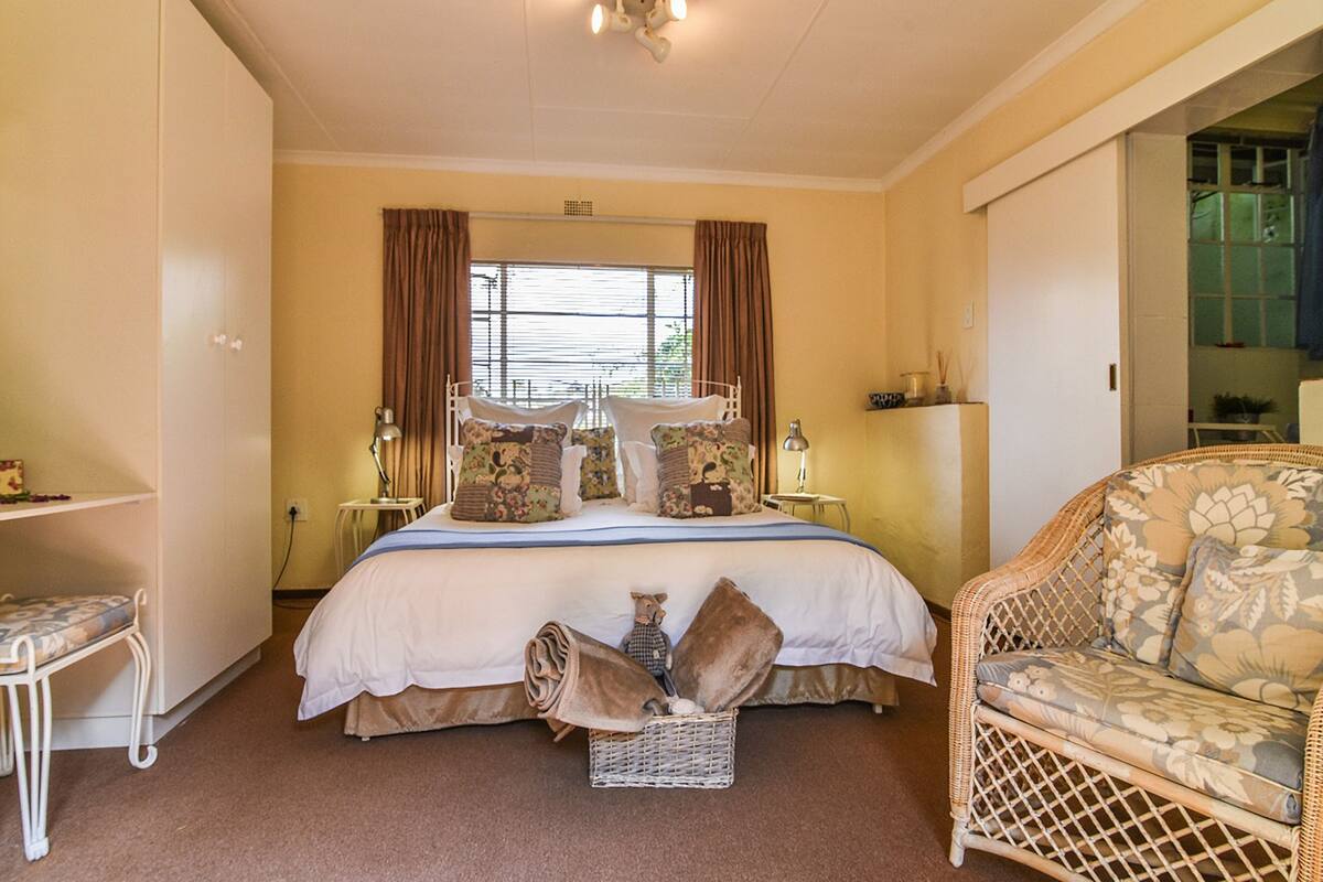 A bedroom with a large bed, a wicker chair, a bedside table, and a window with curtains.