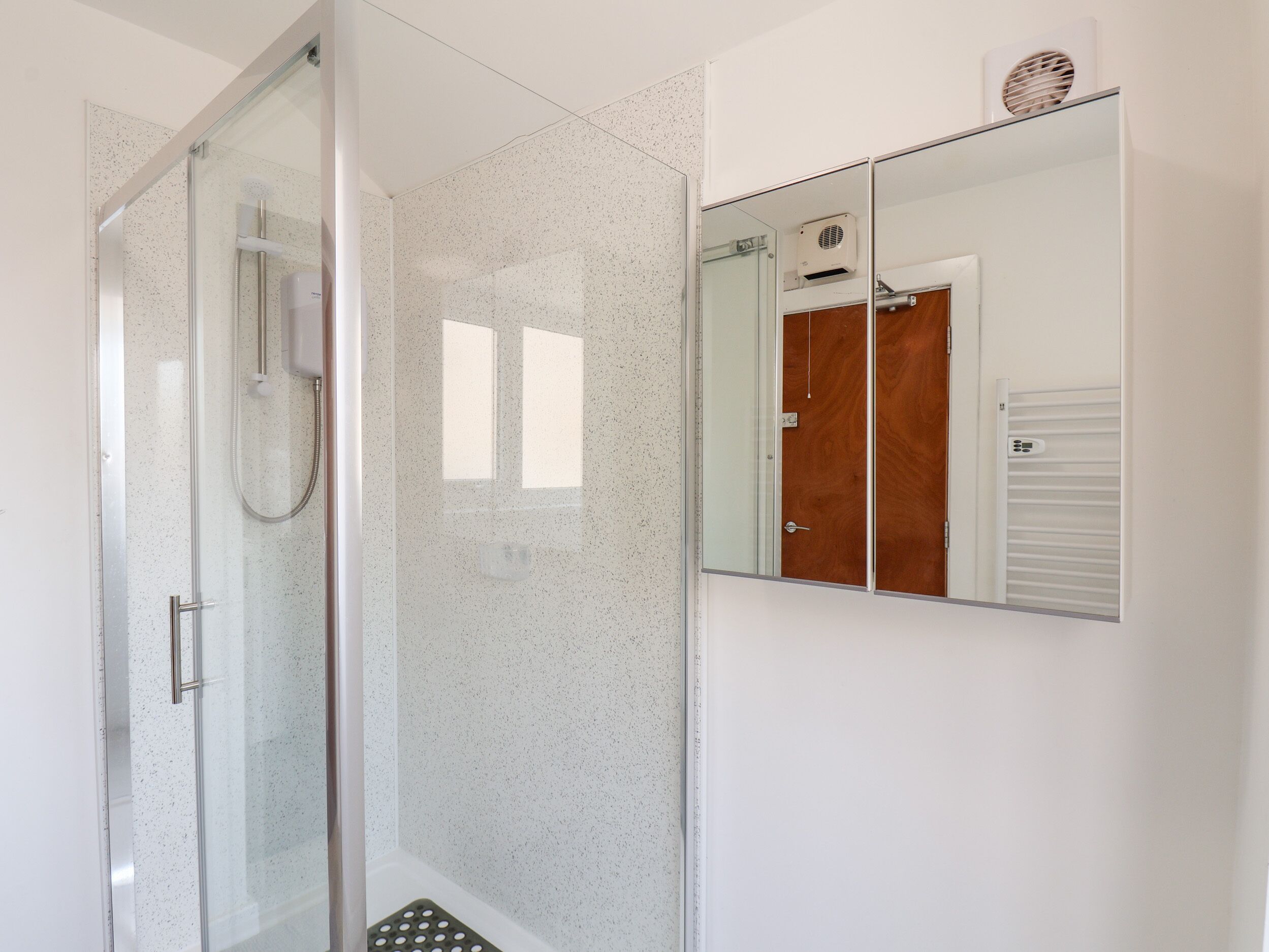 A modern bathroom with a glass shower enclosure, a white speckled wall, and a mirror reflecting a wooden door and a towel rack.