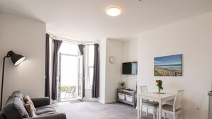 A modern living room with a sofa, a dining table with chairs, a TV, and a view of the outdoors through a large window. - 2 Seaview @ Bridlington Bay (Bridlington)