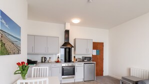 A compact kitchen with white cabinets, a black range hood, a microwave, and a refrigerator. There is a small dining table with a white chair and a vase of red tulips. - 2 Seaview @ Bridlington Bay (Bridlington)