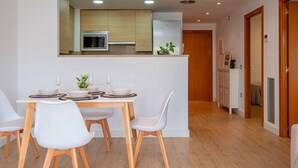 A modern kitchen with a built-in microwave, a dining area with white chairs and a wooden table, and a hallway leading to a bedroom.
