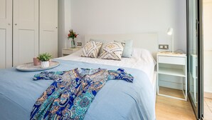 A modern bedroom with a large bed, a bedside table with a lamp, a small potted plant, and a view of the outside through a glass door.