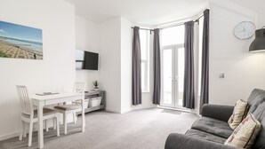 A modern living room with a grey sofa, a white dining table with chairs, a flat-screen TV mounted on the wall, and a large window with grey curtains.