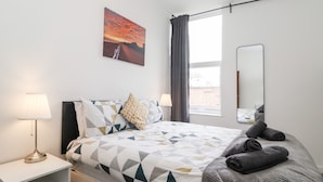 A modern hotel room with a neatly made bed, a bedside lamp, a large window with curtains, and a painting on the wall.