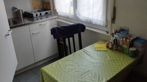 Eine kleine Küche mit weißen Schränken, einer Spüle und einem Fenster mit durchsichtigen Vorhängen. Ein Tisch mit grüner Tischdecke und verschiedenen Gegenständen darauf.