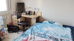 Ein Schlafzimmer mit Bett, Schreibtisch und Stuhl. Außerdem ein Heizkörper, ein Fenster und eine Tür.