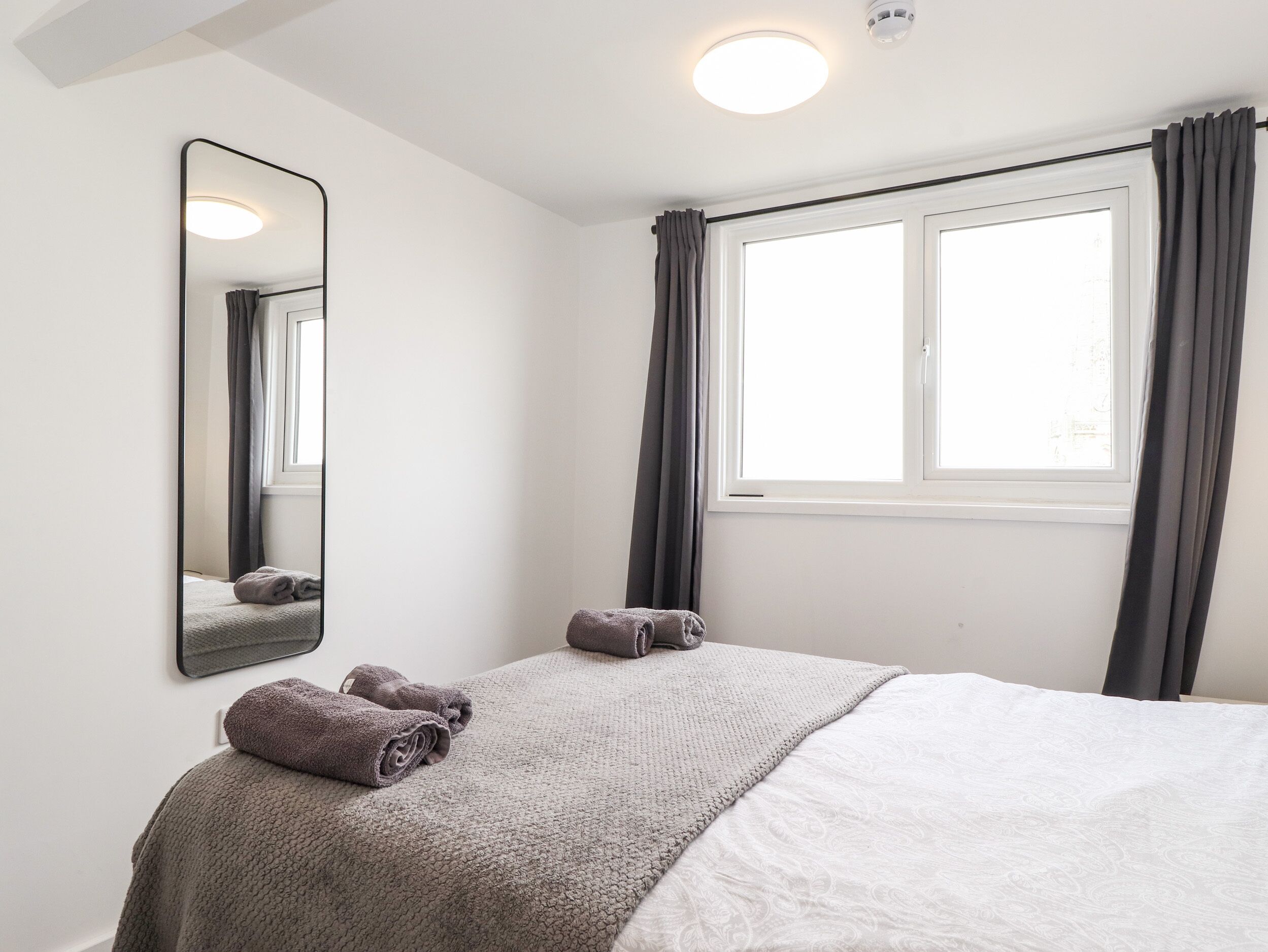 Une chambre à coucher avec un lit, une fenêtre avec des rideaux, un miroir et un luminaire au plafond.